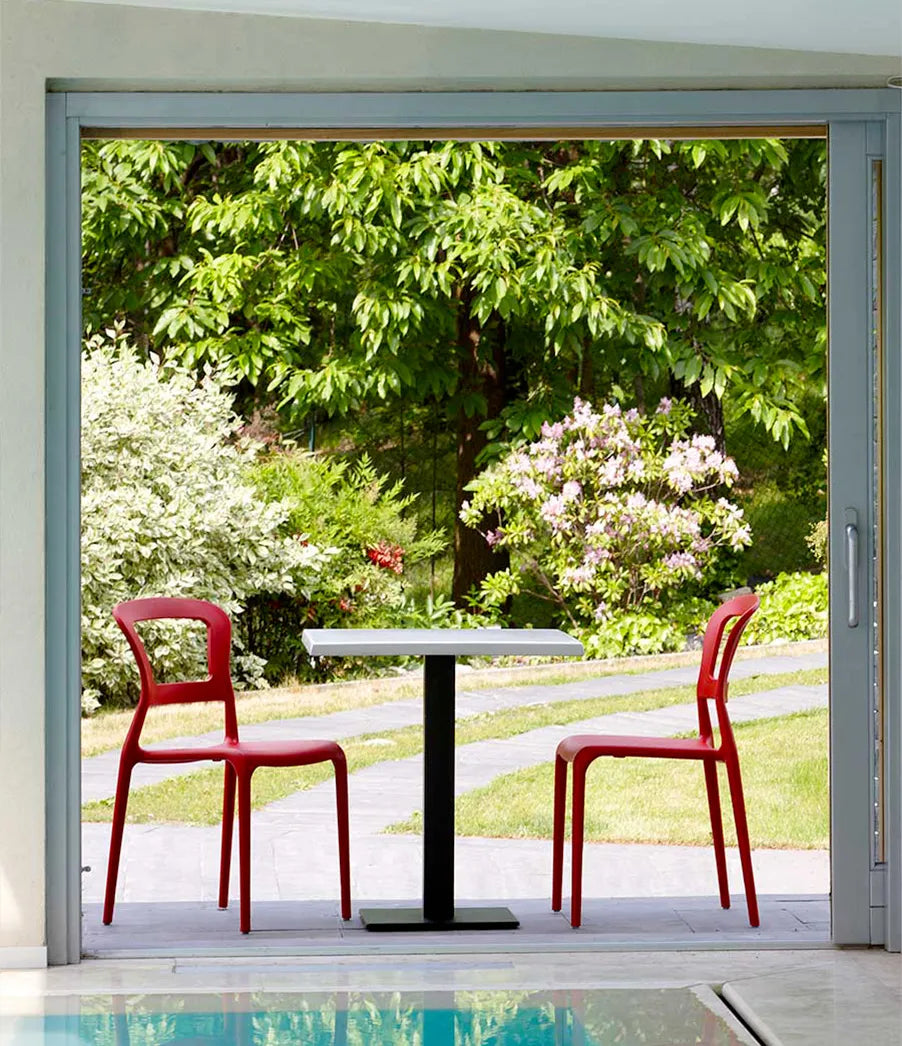 S•CAB Tiffany Bistro Table Base with a square tabletop paired with red Tiffany chairs in a stylish outdoor café setting overlooking a garden.