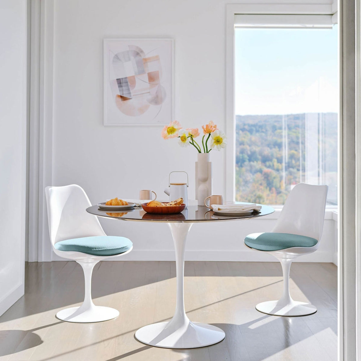 Round Tulip dining table with a white pedestal base paired with white Tulip dining chairs featuring light blue upholstered seat cushions in a bright modern dining room with large windows and natural daylight, inspired by Eero Saarinen's iconic mid-century design