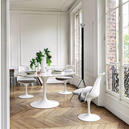 Round Tulip dining table with a white marble top and white pedestal base paired with white Tulip dining chairs featuring grey upholstered seat cushions in an elegant contemporary dining room with parquet flooring and large French windows, inspired by Eero Saarinen's iconic mid-century modern design.